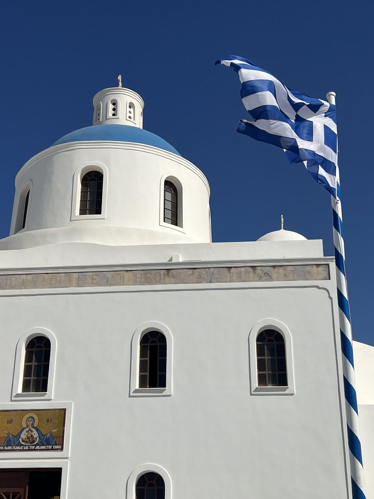 A blue-domed Greek Orthodox church and the Greek flag against a clear sky